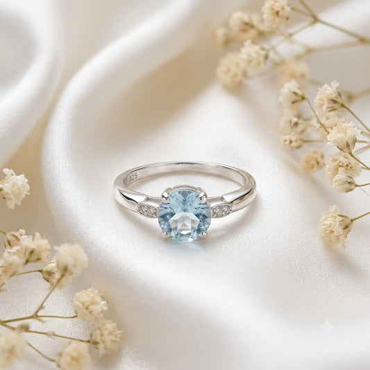Solitaire Blue Topaz Ring crafted in 925 Sterling Silver with a round cut sky blue gemstone and four-prong setting with accent stones on the shoulders.