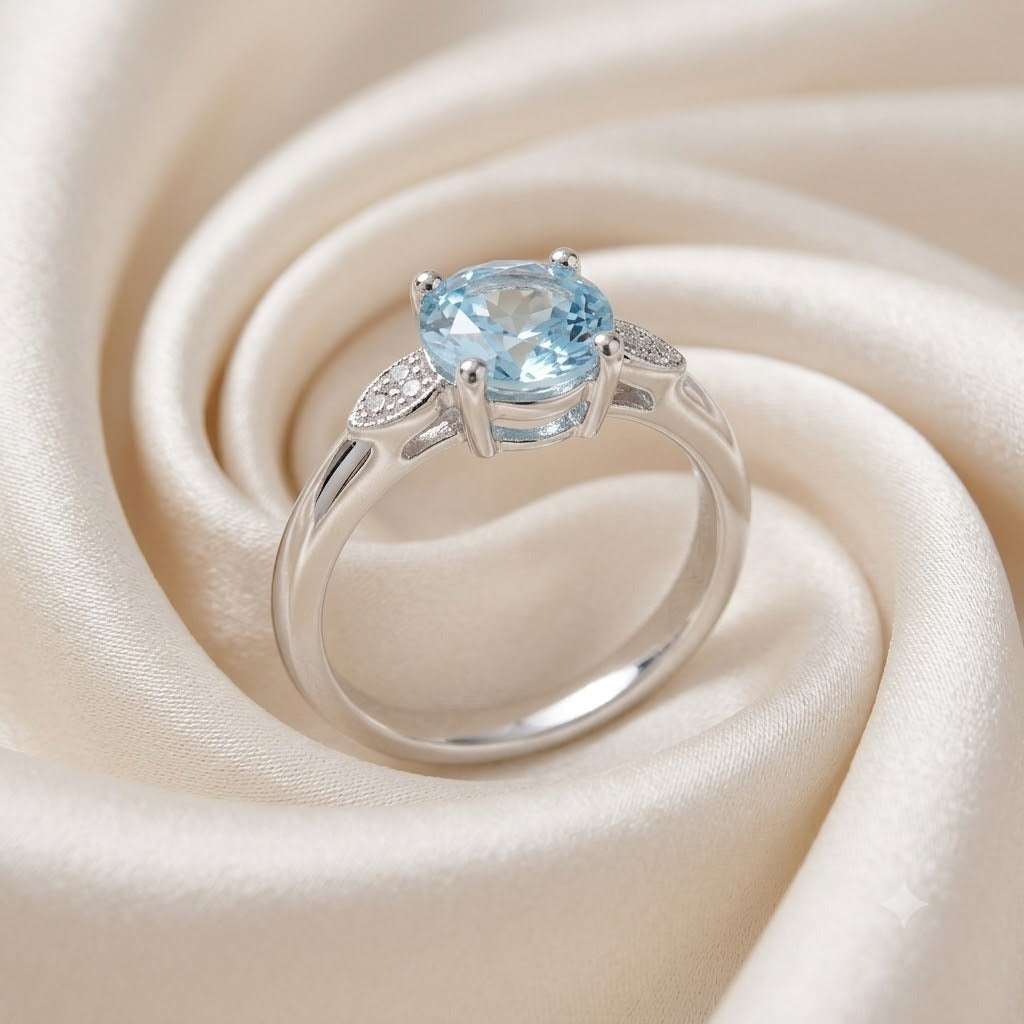 Solitaire Blue Topaz Ring crafted in 925 Sterling Silver with a round cut sky blue gemstone and four-prong setting with accent stones on the shoulders.